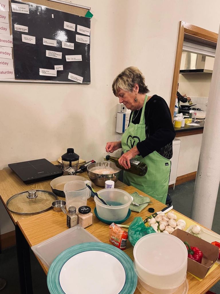 Photo of a woman pouring an oil into a  bowl containing a pale coloured mixture of lentil pancake batter.  Also on the table is an induction hob, various ingredients, containers and cutting boards.